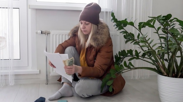 A woman warmly dressed in a brown jacket and hat is sitting near a heating appliance with bills in her hands money and a phone The concept of a crisis and a large set for utilities