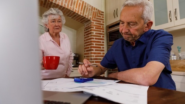 Senior caucasian couple spending time at home together, sitting in kitchen, the man looking at paperwork and the woman holding a cup. isolating during coronavirus covid 19 quarantine lockdown.
