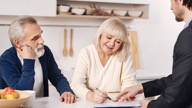 The moment of signing the agreement. Decisive positive senior couple sitting at home and having conversation with social security advisor while signing the agreement