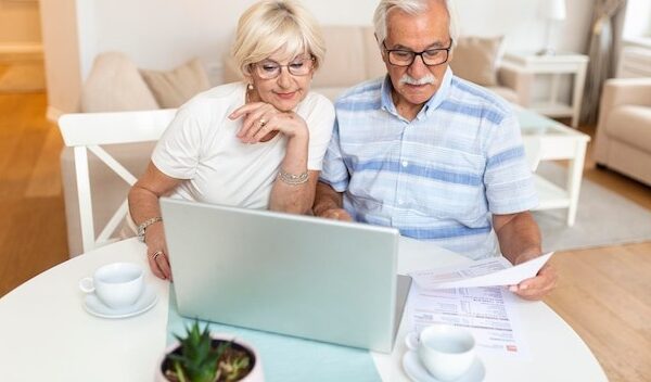 Happy middle aged husband and wife sitting at table with laptop and paper bills calculating domestic incomes together at home