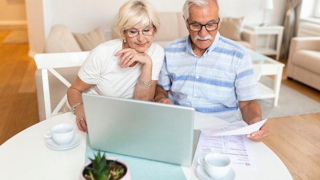 Happy middle aged husband and wife sitting at table with laptop and paper bills calculating domestic incomes together at home
