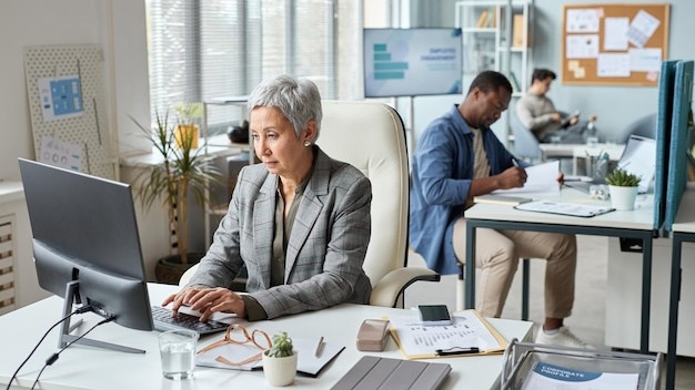 Portrait of senior asian woman working at desk in open office with people in background copy space