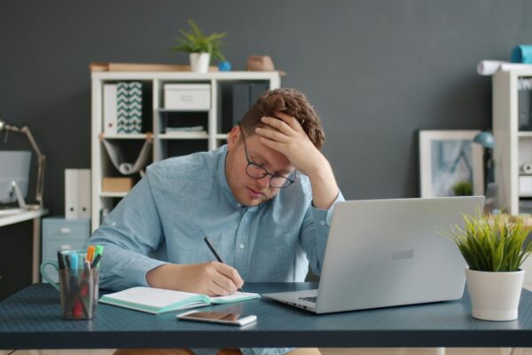 Man writing at desk with laptop, looking stressed.