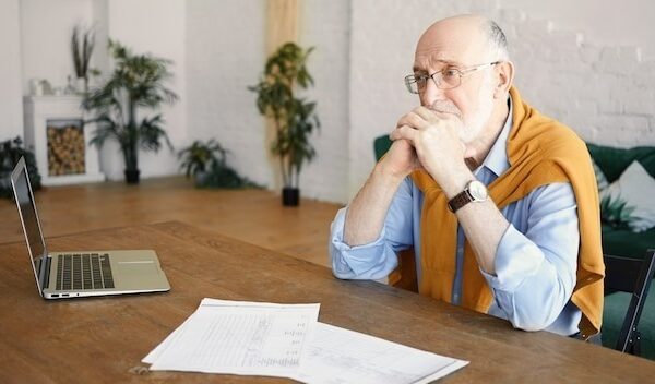 Indoor shot of unhappy sad senior bearded male entrepreneur sitting at desk with laptop and papers having depressed facial expression, frustrated with financial problems, holding hands under his chin