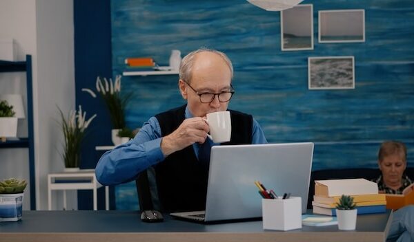 Senior businessman reading reports sitting in front of laptop working from home drinking coffee. Retired man using modern technology analysing typing searching while wife sitting on couch reading a bo