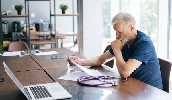 Focused thoughtful gray-haired mature man doctor studying medical report of patient, working with paper documents sitting at desk in office room in medical clinic.