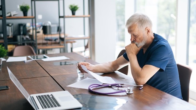 Focused thoughtful gray-haired mature man doctor studying medical report of patient, working with paper documents sitting at desk in office room in medical clinic.