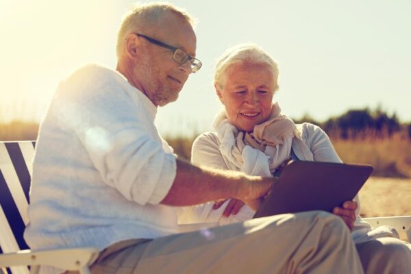 family, age, travel, tourism and people concept – happy senior couple with tablet pc computer resting in folding chairs on summer beach