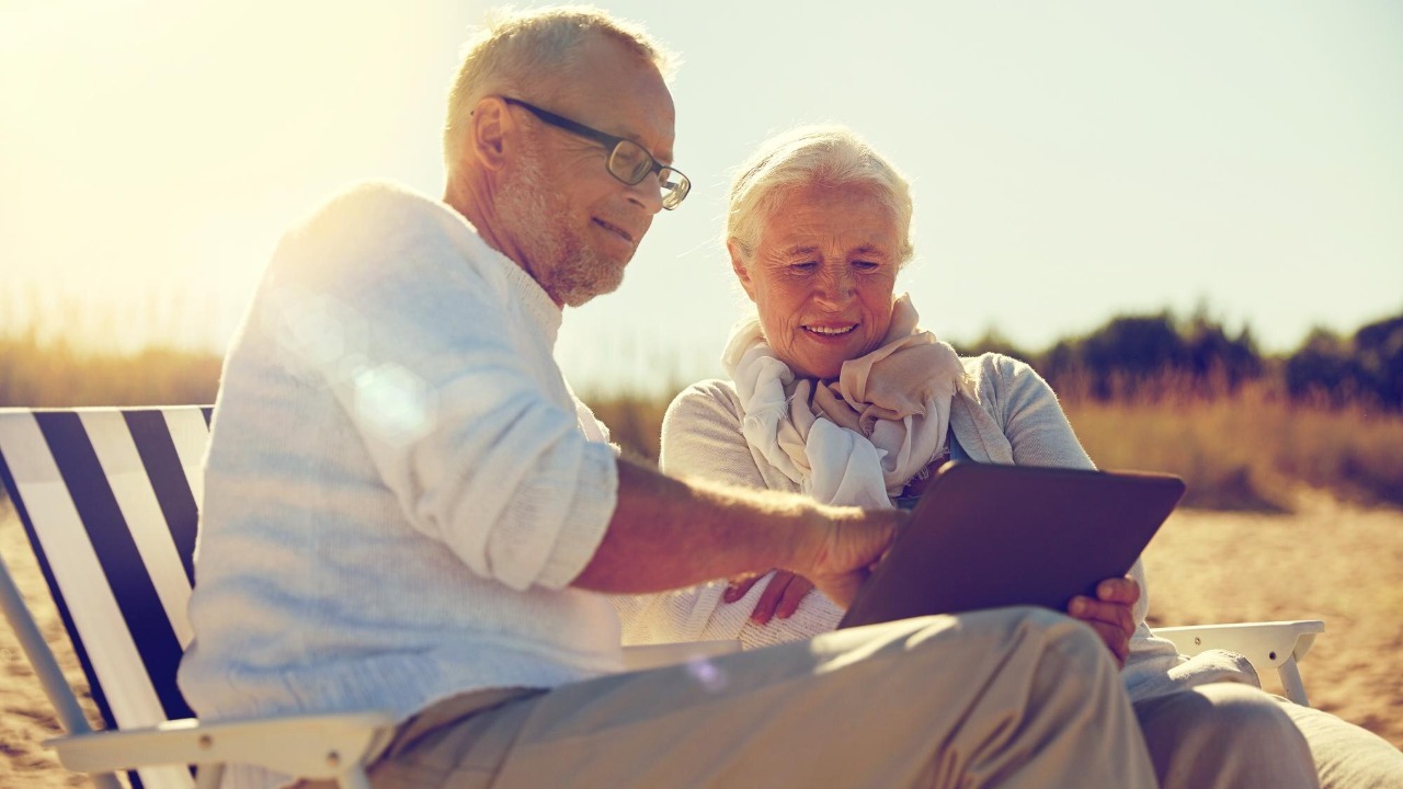 family, age, travel, tourism and people concept – happy senior couple with tablet pc computer resting in folding chairs on summer beach