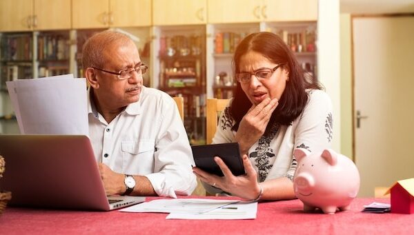 Senior Indian asian couple accounting, doing home finance and checking bills with laptop, calculator and money also with piggy bank while sitting on sofa couch or table at home