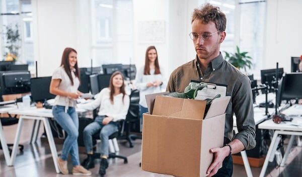 Stylish man in glasses holding box with plant in front of his colleagues in the office.