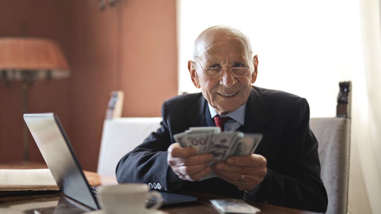 Smiling older man holding a fan of dollar bills at his desk